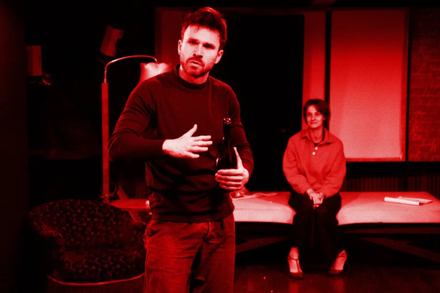 próba do spektaklu „Morderstwo w hotelu", zdjęcie zostało zrobione w sali prób, na zdjęciu: Krzysztof Wach trzymający w ręku butelkę szampana, w tle na łózku siedzi uśmiechnięta Natalia Klepacka, fot. Marcin Nowak. Na zdjęcie został nałożony czerwony filtr.