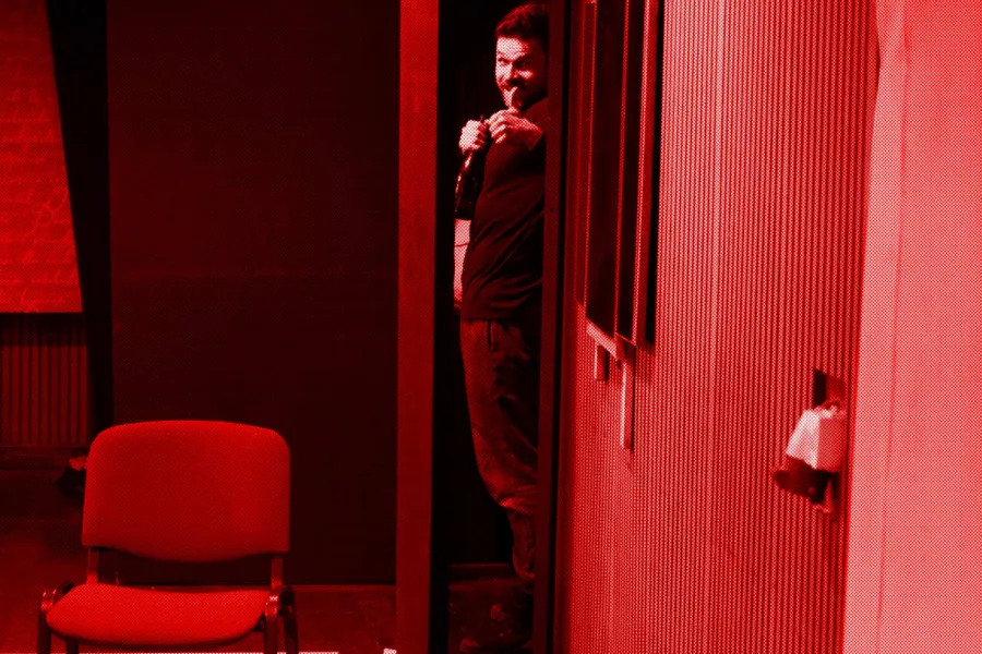 próba do spektaklu „Morderstwo w hotelu", zdjęcie zostało zrobione w sali prób, na zdjęciu: Krzysztof Wach wyglądający zza zastawki, aktor uśmiecha się, w prawej ręce trzyma butelkę szampana, a drugą pokazuje kciuk do góry, fot. Marcin Nowak. Na zdjęcie nałożony jest czerwony filtr.