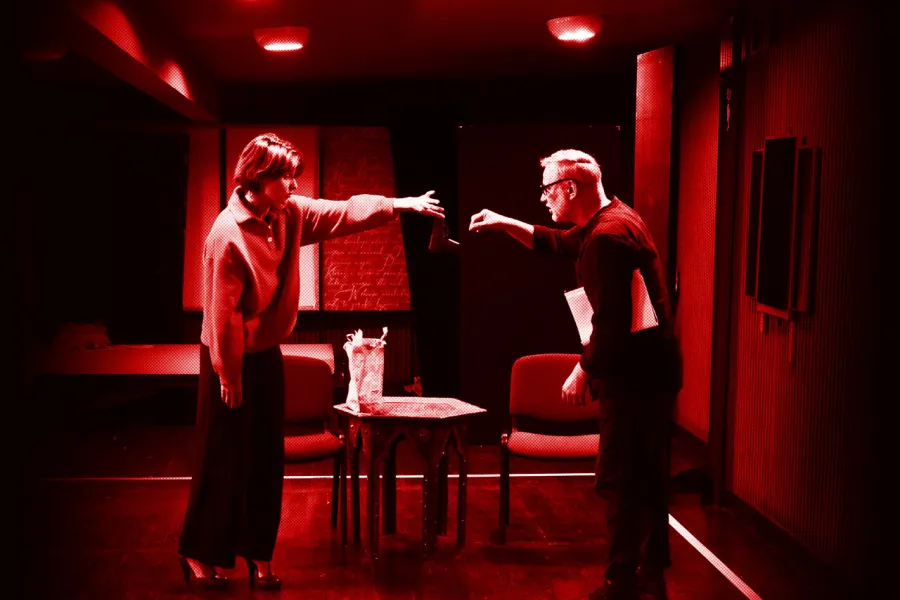 próba do spektaklu „Morderstwo w hotelu", zdjęcie zrobione w sali prób, na zdjęciu: Natalia Klepacka, która w palcach lewej dłoni trzyma pistolet i wręcza go z odległości Mariuszowi Witkowskiemu, fot. Marcin Nowak. Na zdjęcie został nałożony czerwony filtr.