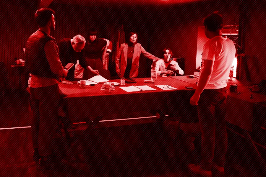 próba spektaklu „Morderstwo w hotelu", zdjęcie zostało zrobione w sali prób, na zdjęciu od lewej, nad długim stołem stoją: Krzysztof Wach, Mariusz Witkowski, inspicjentka Marta Baraszkiewicz, Bogusława Pawelec, Natalia Klepacka. Na przeciwko nich, tyłem do obiektywu stoi Jacek Bończyk - reżyser spektaklu. Fot. Macin Nowak. Na zdjęcie nałożony jest czerwony filtr
