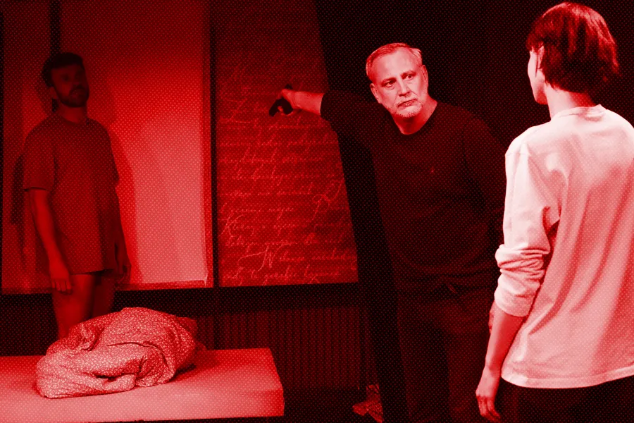 próba spektaklu „Morderstwo w hotelu", zdjęcie zostało zrobione w sali prób, na zdjęciu w tle za łózkiem stoi Krzysztof Wach, przed łózkiem stoi Mariusz Witkowski zwrócony sylwetką w stronę Natalii Klepackiej, ale dłoń z wycelowanym pistoletem ma w kierunku Krzysztofa Wacha, fot. Macin Nowak. Na zdjęcie został nałożony czerwony filtr.