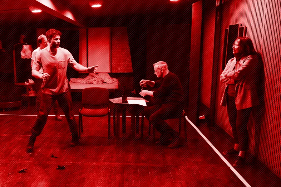 próba spektaklu „Morderstwo w hotelu", zdjęcie zostało wykonane w sali prób, na zdjęciu Krzysztof Wach, z tyłu reżyser Jacek Bończyk, siedzący na krześle przy niskim stole Mariusz Witkowski oraz stojąca przy ścianie Bogusława Pawelec, fot. Macin Nowak. Na zdjęcie nałożony jest czerwony filtr.