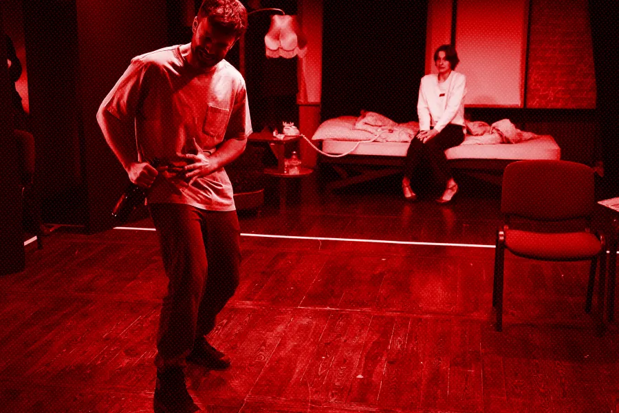 próba spektaklu „Morderstwo w hotelu", zdjęcie zostało wykonane w sali prób, na zdjęciu na pierwszym planie stoi Krzysztof Wach, w ręku ma butelkę szampana, w tle na łóżku siedzi Natalia Klepacka, fot. Macin Nowak. Na zdjęcie nałożony jest czerwony filtr.