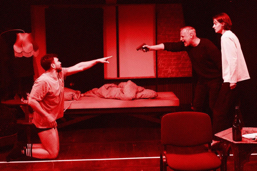 próba spektaklu „Morderstwo w hotelu", zdjęcie zostało wykonane w sali prób, na zdjęciu Krzysztof Wach, który ma na sobie koszulkę i bokserki, klęczy i wskazuje palcem na Mariusza Witkowskiego, Mariusz Witkowski celuje pistoletem do Krzysztofa Wacha, przy nim stoi Natalia Klepacka. fot. Macin Nowak. Na zdjęcie został nałożony czerwony filtr.