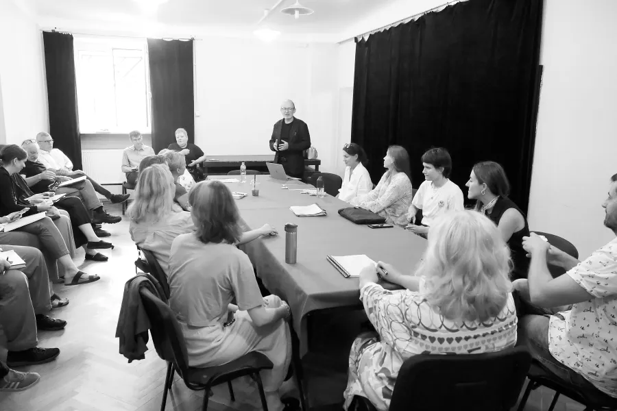 pierwsza próba spektaklu „Czas na wielki numer". Zdjęcie zostało zrobione na sali prób. Po środku widać duży stół nakryty suknem, na stole znajdują się egzemplarze scenariusza, notesy oraz kubki. Przy stole siedzą realizatorzy i aktorzy występujący w spektaklu. Na końcu stołu po prawej stronie stoi dyrektor teatru Paweł Łysak. Wokół stołu na krzesłach siedzą pracownicy teatru. Na zdjęcie nałożony jest czarno-biały filtr. Fot. Marcin Nowak