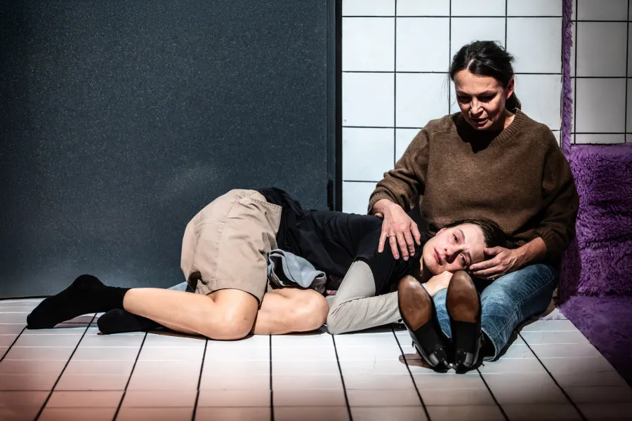 Zdjęcie ze spektaklu „Czas na wielki numer". Na fotografii widać aktorkę Natalię Klepacką, która leży na białych kafelkach i trzyma głowę na kolanach aktorki Moniki Badowskiej. Natalia Klepacka ubrana jest w krótkie beżowe, luźne spodnie i czarną koszulkę z długim, szarym rękawem. Na stopach ma czarne skarpetki.  Monika Badowska głaszcze Natalię Klepacką. Jest ubrana w niebieskie jeansy i brązowy sweter. Autorką zdjęcia jest Natalia Kabanow