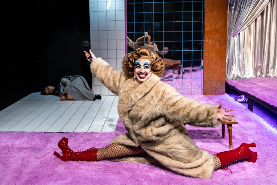 Zdjęcie ze spektaklu „Czas na wielki numer". Na fotorafii widać aktorkę Matyldę Paszczenko w charakteryzacji na drag queen. Ma na sobie gruby płaszcz. Robi szpagat. W tle pod drzwiami leży aktorka Natalia Klepacka. Autorką zdjęcią jest Natalia Kabanow