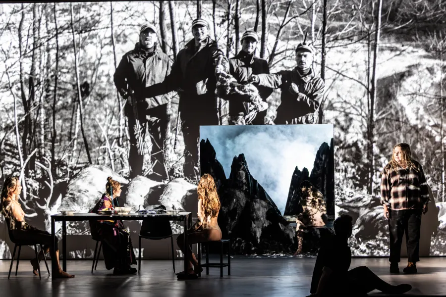 Zdjęcie ze spektaklu „Prowadź swój pług przez kości umarłych". Na Dużej Scenie Teatr Jaracza widać długi stół przy którym siedzą dwie tancerki i aktorka Sara Lityńska, patrzą się na wyświetlane na ścianie zdjęcie myśliwych, w ścianie jest kwadratowa dziura przez którą widać góry. Obok dziury stoi aktorka Milena Lisiecka. Autorką zdjęcia jest Natalia Kabanow