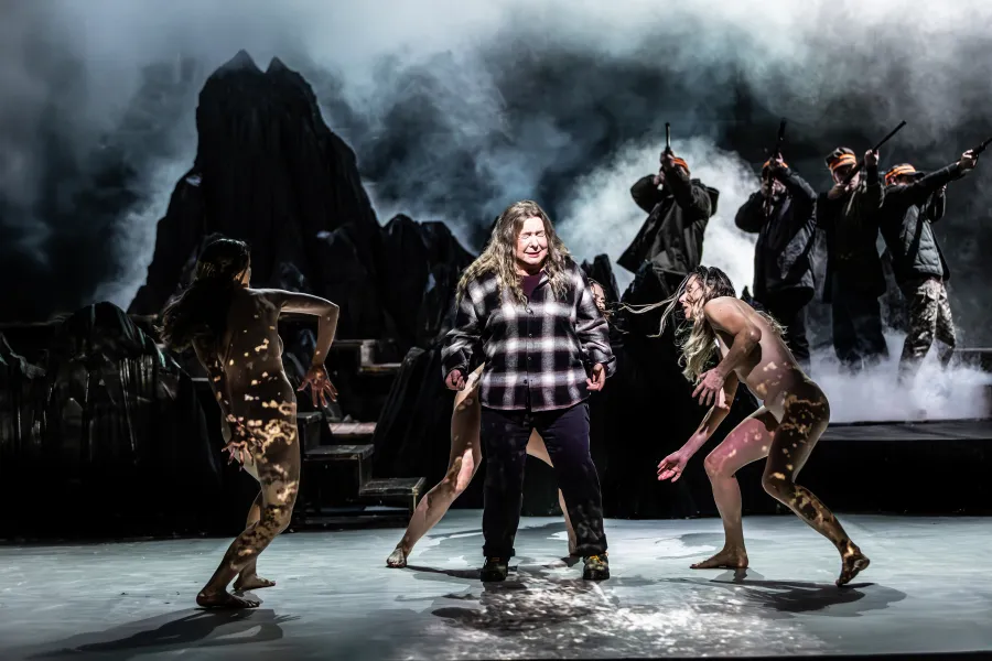 Zdjęcie ze spektaklu „Prowadź swój pług przez kości umarłych". Na scenie po środku stoi aktorka Milena Lisiecka, z grymasem płaczu na twarzy. Wokół niej tańczą trzy tancerki odgrywające role saren. Za nimi, wśród scenografii imitującej góry, stoi czwórka aktorów grających myśliwych, celującycyh w cel strzelbami. 