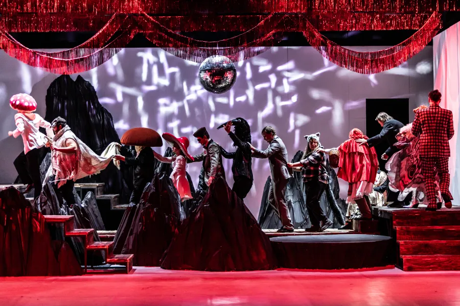 Zdjęcie ze spektaklu „Prowadź swój pług przez kości umarłych" w reżyserii Leny Frankiewicz, na scenie widzimy dwunastu aktorów w senografii przypominającej góry. Każdy z bohaterów ma przebranie. Nad sceną wisi kula dyskotekowa. Jest czerwone światło na scenie. Jest to scena balu grzybiarza. Autorką fotografii jest Natalia Kabanow