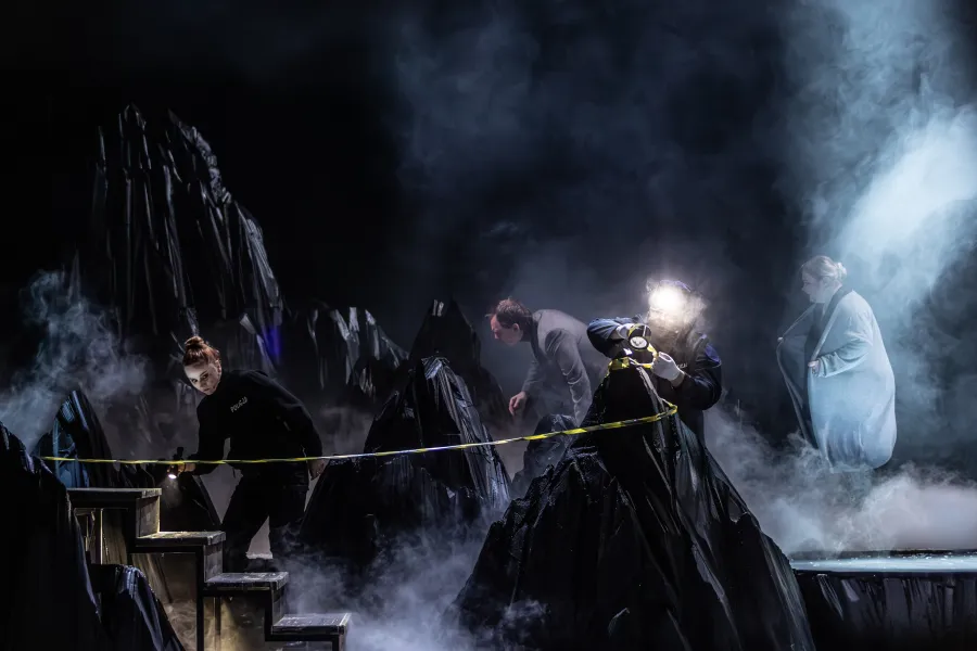 Zdjęcie ze spektaklu „Prowadź swój pług przez kości umarłych". Na zdjęciu widać 4 aktorów, którzy odgrywają scenę śledztwa. Dwie osoby w kostiumach policjantów świecą latarkami. Z tyłu stoi kobieta w jasnym kardiganie. Na scenie jest ciemno, jest bardzo dużo gęstego dymu. Autorką zdjęcia jest Natalia Kabanow