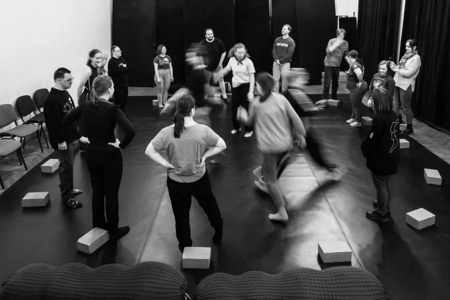 Czarno-białe zdjęcie przedstawiające grupę osób w sali prób. Uczestnicy są ustawieni w szerokim kręgu na ciemnej podłodze, a przed wieloma z nich leżą jasne, prostokątne klocki. Część osób stoi nieruchomo, obserwując środek kręgu, gdzie dwie postacie są rozmyte w ruchu, co sugeruje dynamikę ćwiczenia lub zabawy ruchowej. W tle widać proste ściany, rzędy krzeseł oraz ciemne kotary. Atmosfera wydaje się skupiona i warsztatowa.