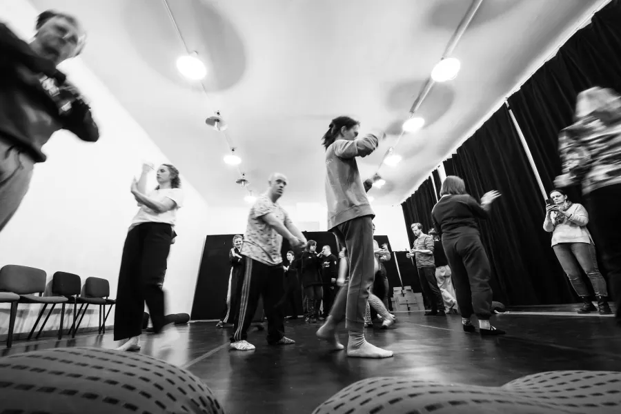 Czarno-białe zdjęcie wykonane z niskiej perspektywy (z poziomu podłogi), przedstawiające grupę osób podczas dynamicznych warsztatów ruchowych w sali z ciemną podłogą i białym sufitem. Wiele postaci jest rozmytych, co podkreśla ekspresję i tempo ćwiczeń. Uczestnicy wykonują różnorodne gesty rękami i są w trakcie przemieszczania się po przestrzeni. Na suficie widać rzędy jasnych, okrągłych lamp, a w tle czarną kotarę i rządy krzeseł. Na pierwszym planie widoczne są fragmenty wypukłych, fakturowanych puf.