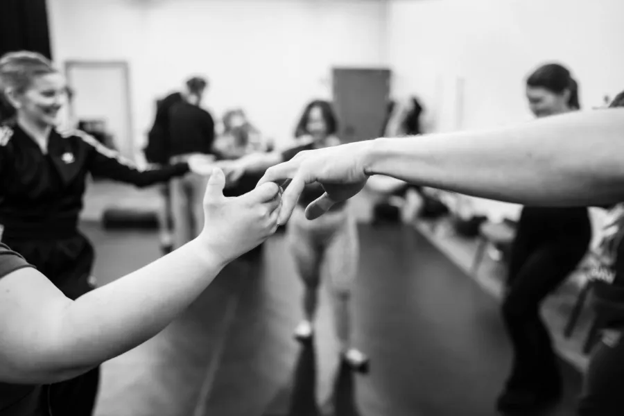 Czarno-białe zdjęcie o płytkiej głębi ostrości, przedstawiające zbliżenie na dłonie dwóch osób, które delikatnie stykają się palcami w geście porozumienia lub wspólnego ruchu. Punkt ostrości jest ustawiony na dłoniach znajdujących się w centrum kadru. W rozmazanym, jasnym tle widać sylwetki pozostałych uczestników warsztatów, w tym uśmiechniętą kobietę po lewej stronie. Scena oddaje atmosferę bliskości, zaufania i emocjonalnego zaangażowania podczas zajęć grupowych w sali prób.