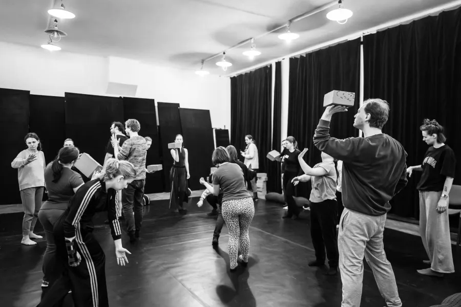 Czarno-białe zdjęcie przedstawiające grupę osób podczas ćwiczeń warsztatowych w sali z ciemną podłogą i czarnymi kotarami. Uczestnicy wchodzą w interakcje z jasnymi, prostokątnymi klockami – niektórzy trzymają je w dłoniach, inni balansują nimi na głowie lub ramieniu, a jeszcze inni wykonują gesty bez klocków. Na pierwszym planie po prawej stronie mężczyzna stoi profilem, trzymając klocek na dłoni przy głowie. Po lewej stronie kobieta w dresie z paskami jest pochylona w dynamicznej pozie.