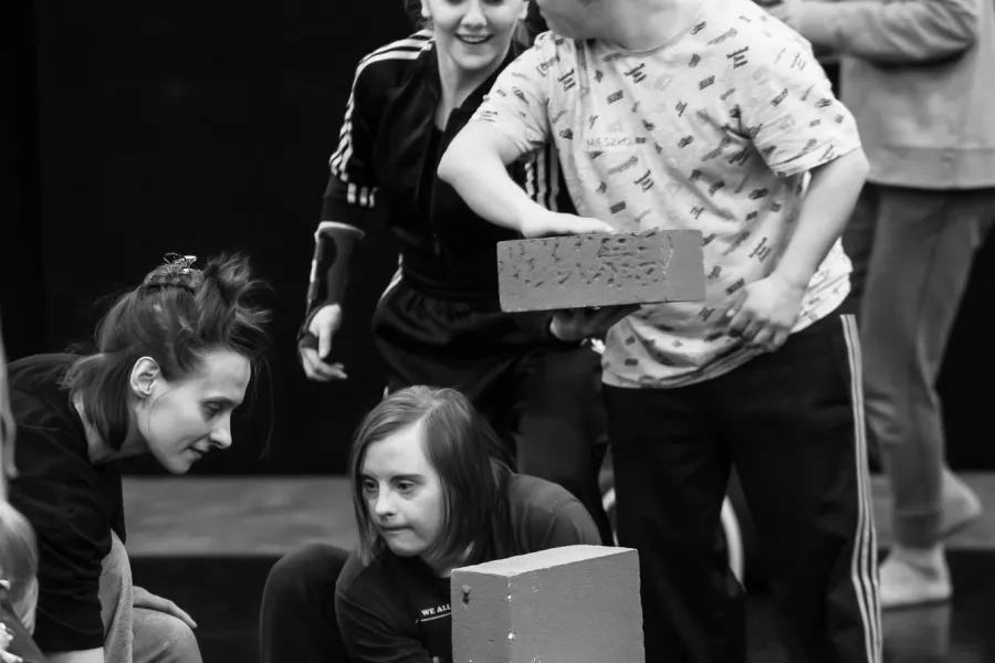 Czarno-białe zdjęcie w pionowym kadrze, przedstawiające grupę osób wspólnie budujących konstrukcję z jasnych, prostokątnych klocków na ciemnej podłodze. W centrum uwagi znajduje się mężczyzna w jasnej koszulce, który precyzyjnie dokłada klocek na szczyt wieży. Obok niego uśmiechnięta kobieta w sportowej bluzie z uwagą śledzi jego ruchy. Dwie inne osoby kucają przy podstawie budowli, a jedna z nich wyciąga dłoń, by podtrzymać lub skorygować ustawienie dolnego elementu.