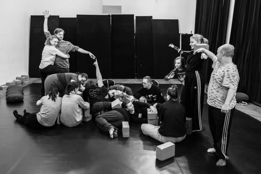 Czarno-białe zdjęcie panoramiczne przedstawiające grupę osób tworzącą wspólną, wielopoziomową rzeźbę z własnych ciał i jasnych klocków na środku sali. Część osób leży lub siedzi na podłodze, podczas gdy inne stoją, tworząc wyższe punkty kompozycji. Po lewej stronie mężczyzna i kobieta splatają ramiona, unosząc dłonie w górę. Po prawej stronie uczestnicy balansują klockami na barkach i wyciągają ręce w bok. Całość tworzy dynamiczną, geometryczną strukturę na tle czarnych paneli i kotar.